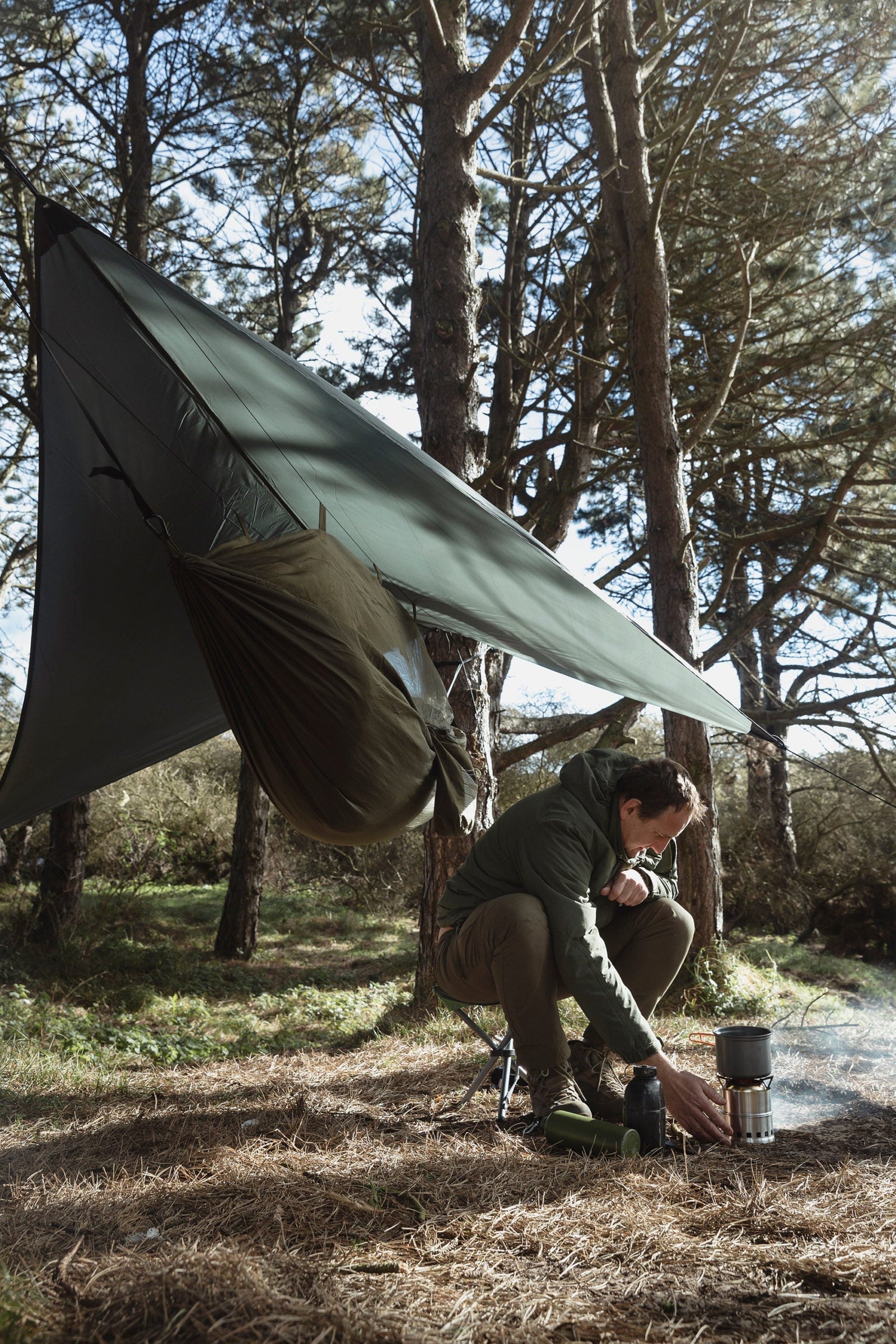 Eine Person in Outdoor-Ausrüstung bereitet mit einem tragbaren Kocher unter einer Plane im Wald Essen zu. In der Nähe hängt eine HIGHLANDER FORCES Highlander Hängematte Crusader Waterproof Hammock, während das Sonnenlicht durch die hohen Bäume des Campingplatzes fällt.
