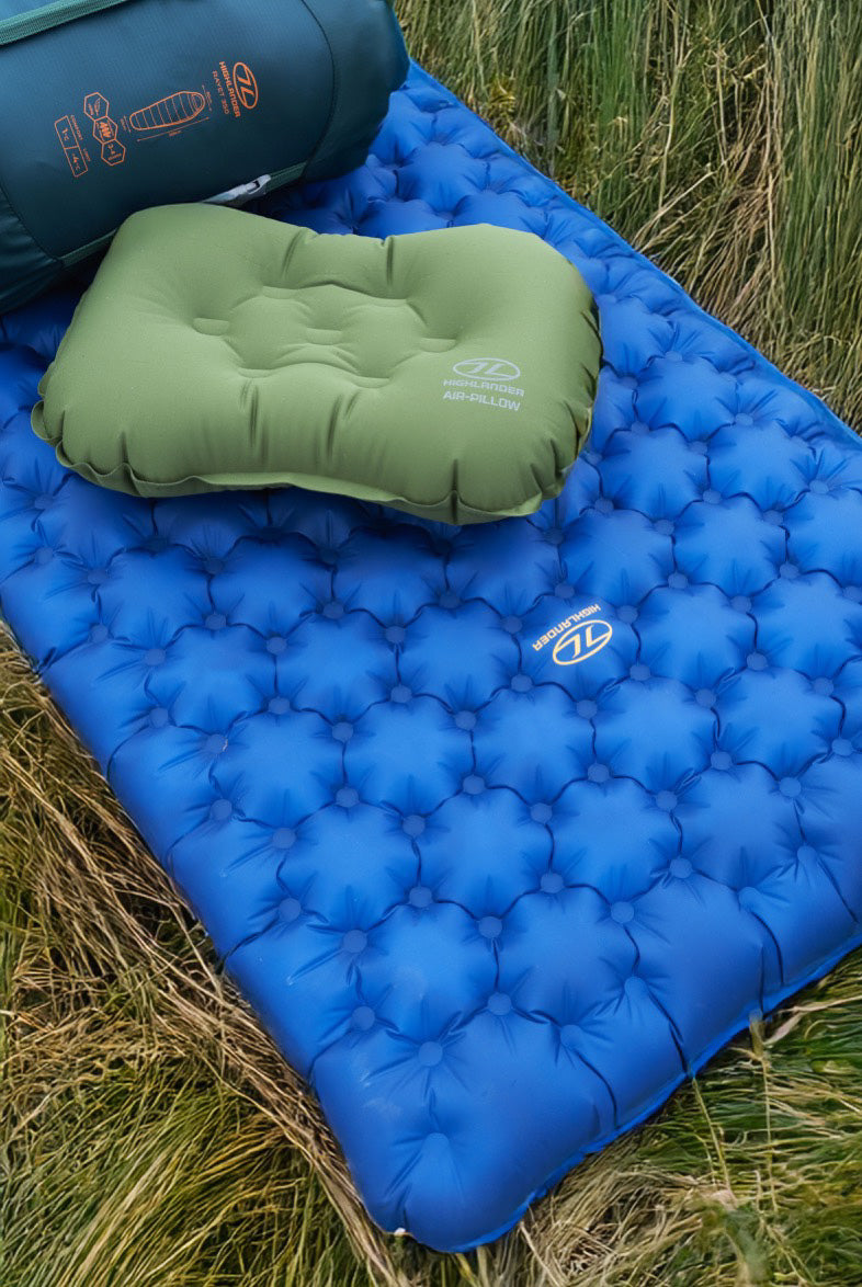 Ein grünes aufblasbares Kissen und eine blaue Highlander NAP PAK ARCTIC AIR MAT von HIGHLANDER FORCES, beide mit Markenlogos versehen, liegen im Freien auf hohem Gras, was an eine Campingszene erinnert.