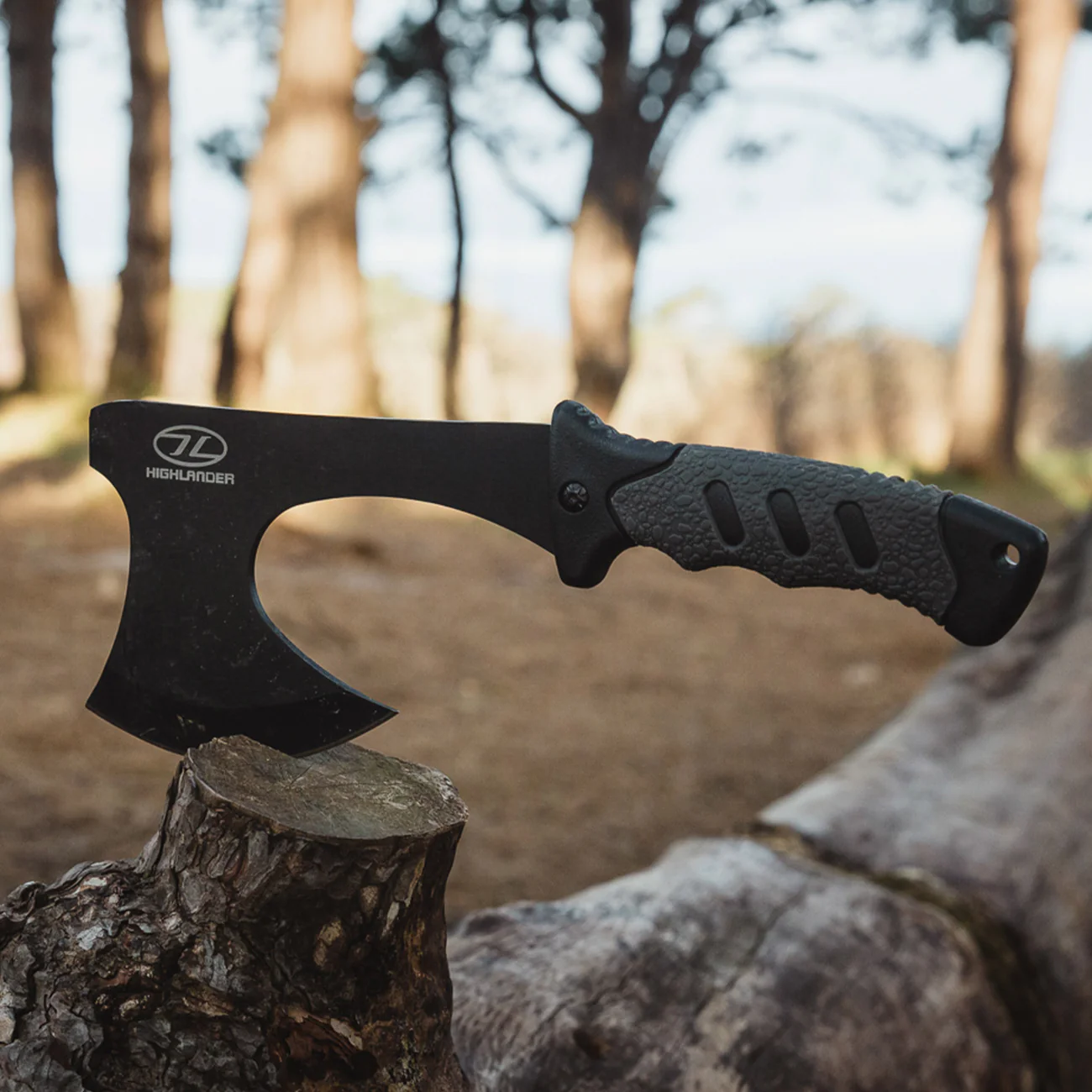 Eine Highlander Survival Axt mit schwarzem Griff von HIGHLANDER FORCES steckt in einem Baumstumpf im Freien, mit hohen Bäumen und sanftem Sonnenlicht im Hintergrund.