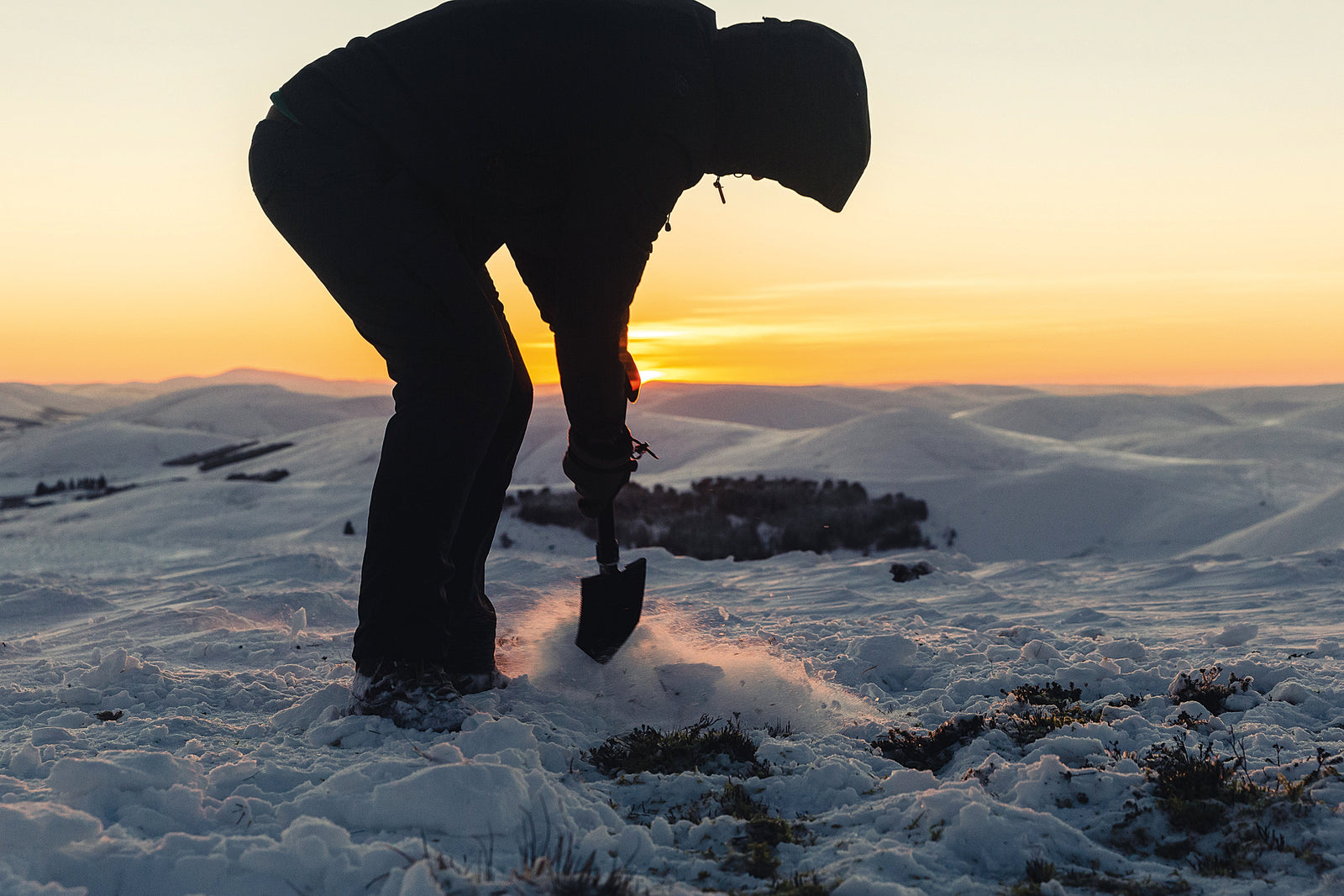 Bei Sonnenuntergang gräbt eine Person in dunkler Winterkleidung mit der Highlander doppelt klappbare Schaufel von HIGHLANDER FORCES inmitten verschneiter Hügel in den Schnee, ausgestattet mit unverzichtbarer Überlebensausrüstung fürs Camping.