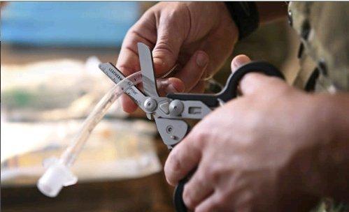 Eine Person benutzt einen LEATHERMAN Schere Raptor, um einen durchsichtigen medizinischen Schlauch zu schneiden, wahrscheinlich während eines Notfalls. Die Hände und das Werkzeug sind vor einem unscharfen Hintergrund im Fokus.