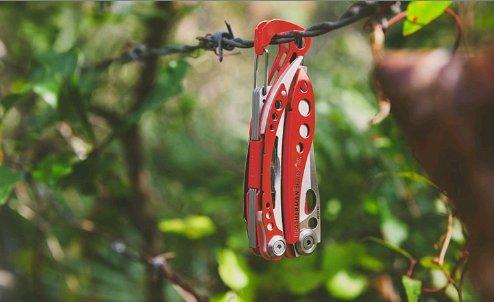 Ein LEATHERMAN Skeletool RX Multitool in Rot und Silber hängt an einem Stacheldraht im Freien, mit verschwommenem grünen Laub im Hintergrund.