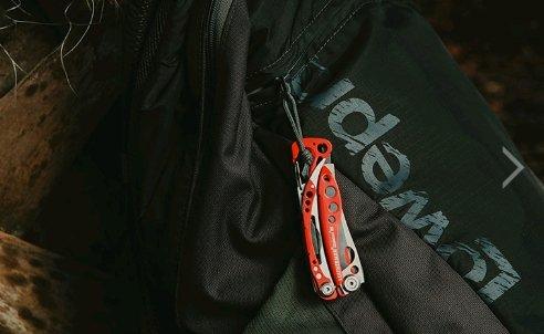 Ein rotes LEATHERMAN Skeletool RX Multitool ist am Reißverschluss eines schwarzen Rucksacks befestigt, wobei der Stoff und das Logo des Rucksacks teilweise sichtbar sind.