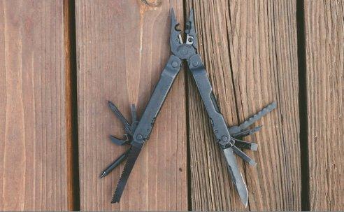 Ein LEATHERMAN Super Tool 300 EOD BLACK Multitool mit einer erweiterten Zange, einer Säge und einem Messer wird auf einer Holzoberfläche mit sichtbarer Maserung präsentiert.