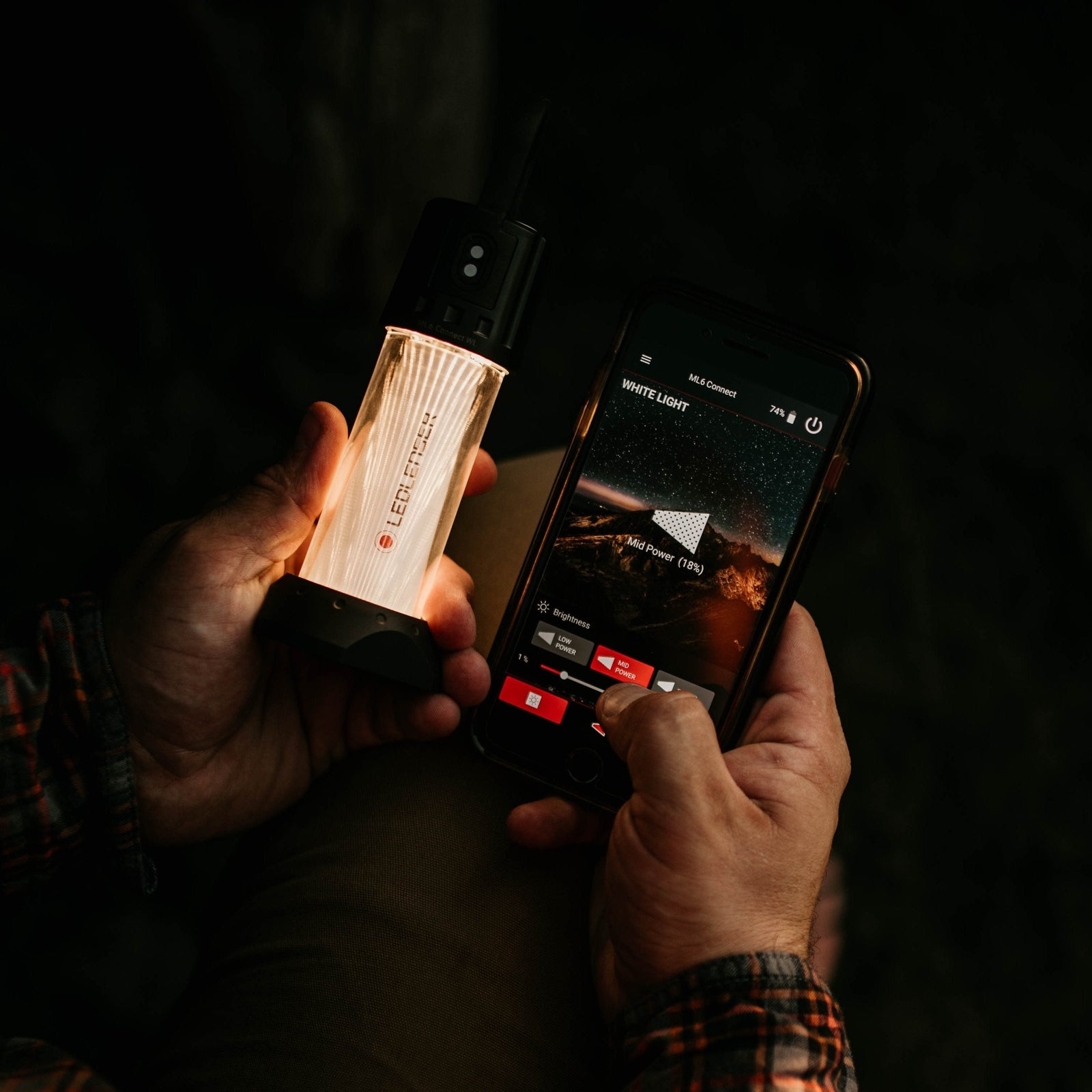Eine Person hält eine LEDLENSER Campinglampe ML6 Connect WL in der einen Hand und steuert mit dem Smartphone in der anderen Hand ihr warmweißes Licht über eine App. Die gedämpfte Einstellung empfiehlt sich bei Nacht oder schlechten Lichtverhältnissen.