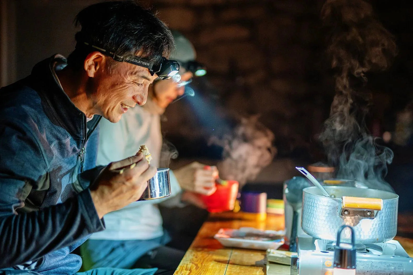 Ein lächelnder Mann mit einer LEDLENSER Ledlenser HF6R Core Stirnlampe (800 Lumen) isst aus einer Metalltasse an einem dampfenden Campingkocher an einem dunklen Holztisch, während eine andere Person im Hintergrund ebenfalls eine Stirnlampe trägt.