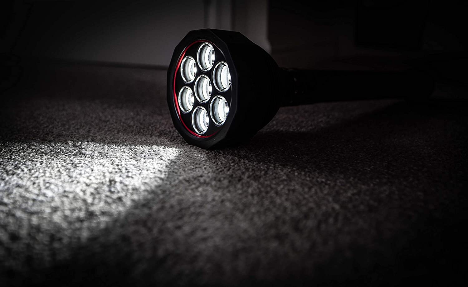 Eine schwarze LEDLENSER Lampe X21R mit sechs runden LEDs liegt auf einem grauen Teppich und wirft einen hellen Lichtstrahl über den Boden in einem dunklen Raum.