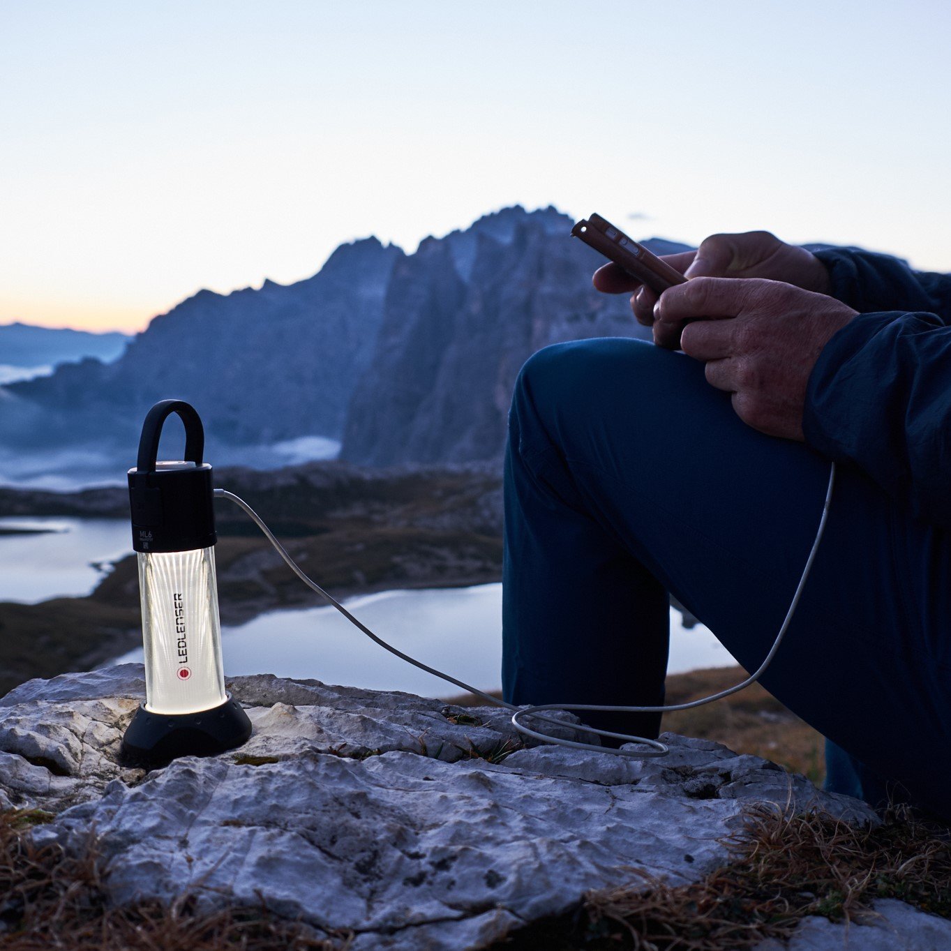 In der Abenddämmerung an einem See und in den Bergen lädt jemand sein Handy mit einer LEDLENSER ML6 Outdoor-Campinglampe auf einem Felsen auf und genießt die raue Naturlandschaft.