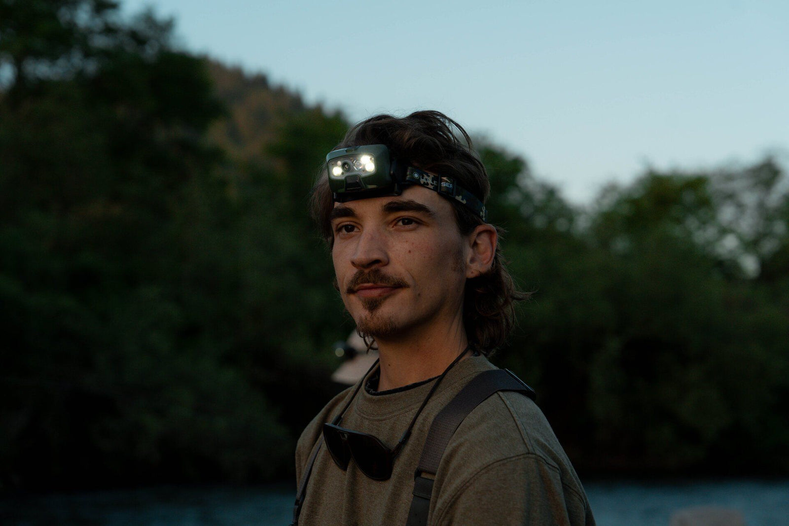 Ein Mann mit Schnurrbart, grünem Hemd und Sonnenbrille steht in der Dämmerung im Freien in der Nähe von Bäumen. Seine LEDLENSER Strinlampe HF8R Core RGB wirft einen adaptiven Lichtstrahl auf die Szene.