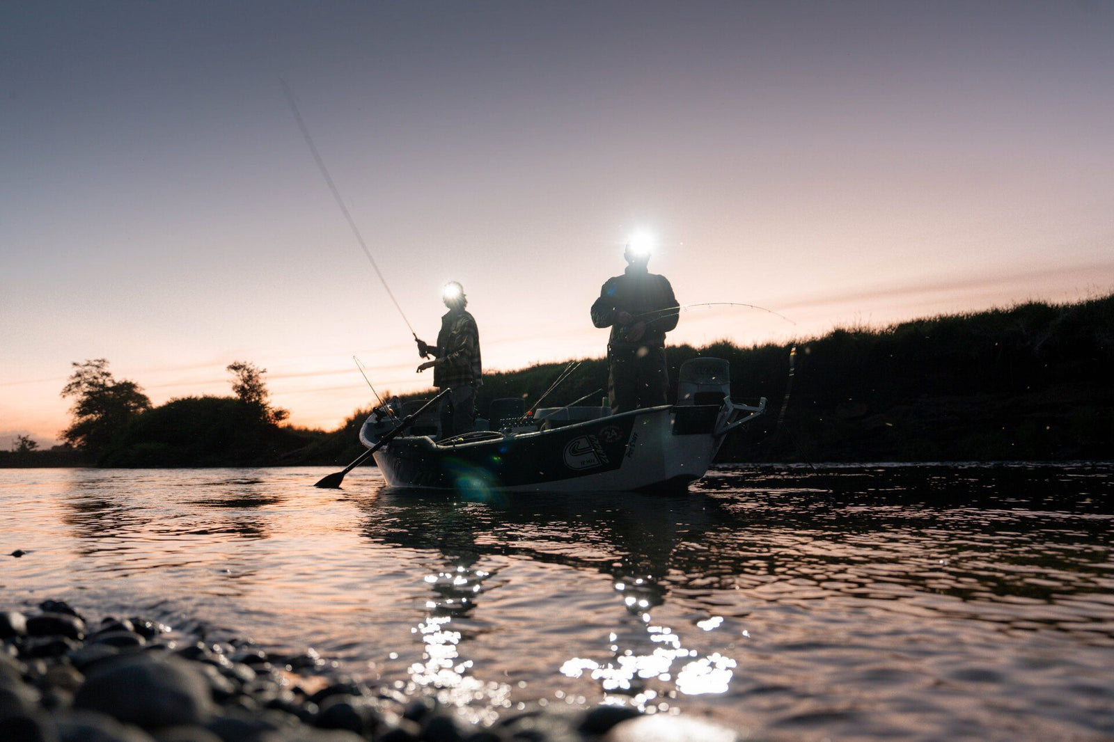 Zwei Personen mit LEDLENSER Strinlampe HF8R Core RGB-Scheinwerfern angeln in der Abenddämmerung auf einem kleinen Boot. Wasser und Ufer sind sichtbar, während ein adaptiver Lichtstrahl der Scheinwerfer den Himmel sanft beleuchtet.