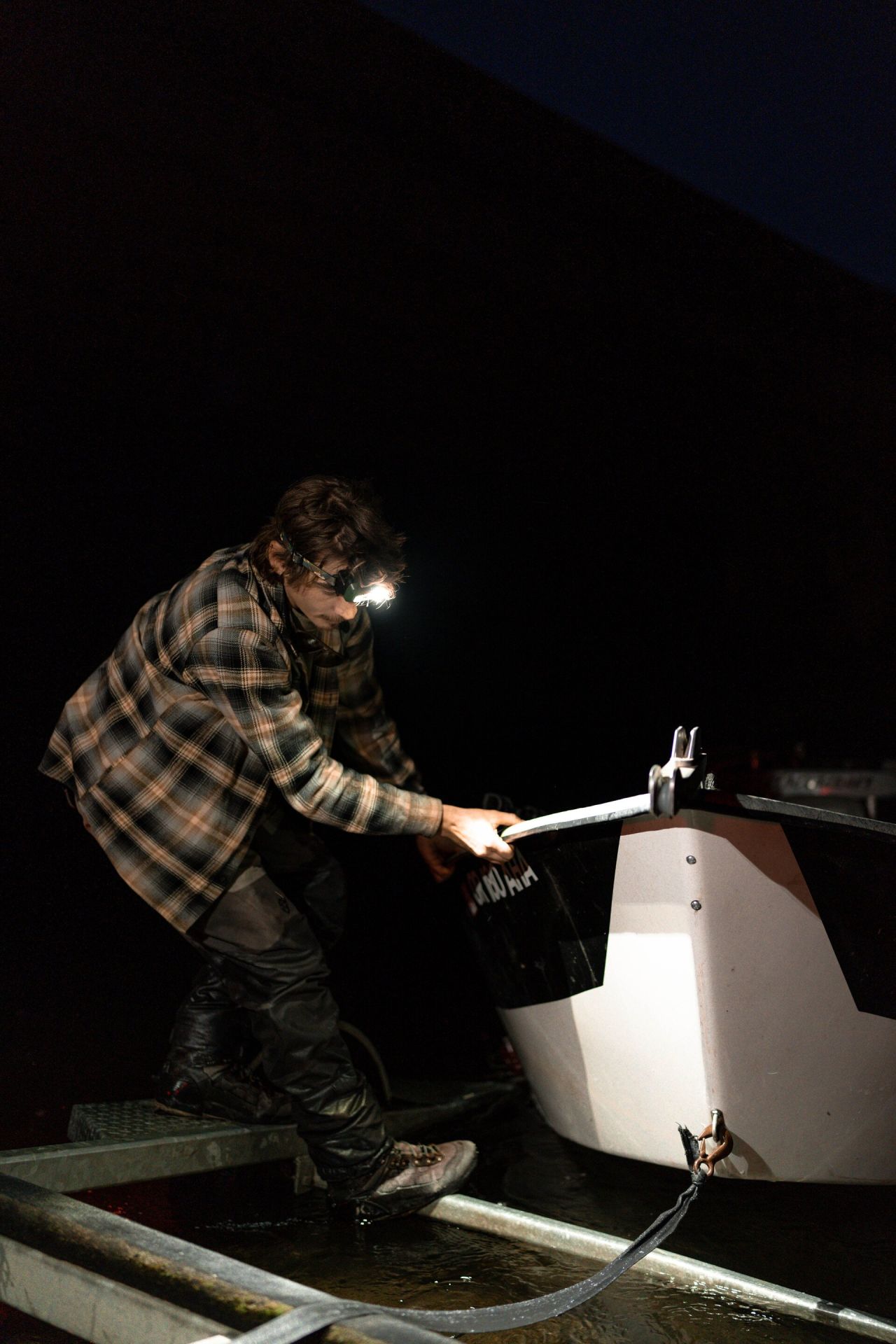 Eine Person in einem karierten Hemd, die die LEDLENSER Strinlampe HF8R Core RGB trägt, arbeitet nachts und sichert ein weißes Boot auf einem Anhänger. Das gedämpfte Licht hebt ihren konzentrierten Gesichtsausdruck und den Bug des Bootes hervor.