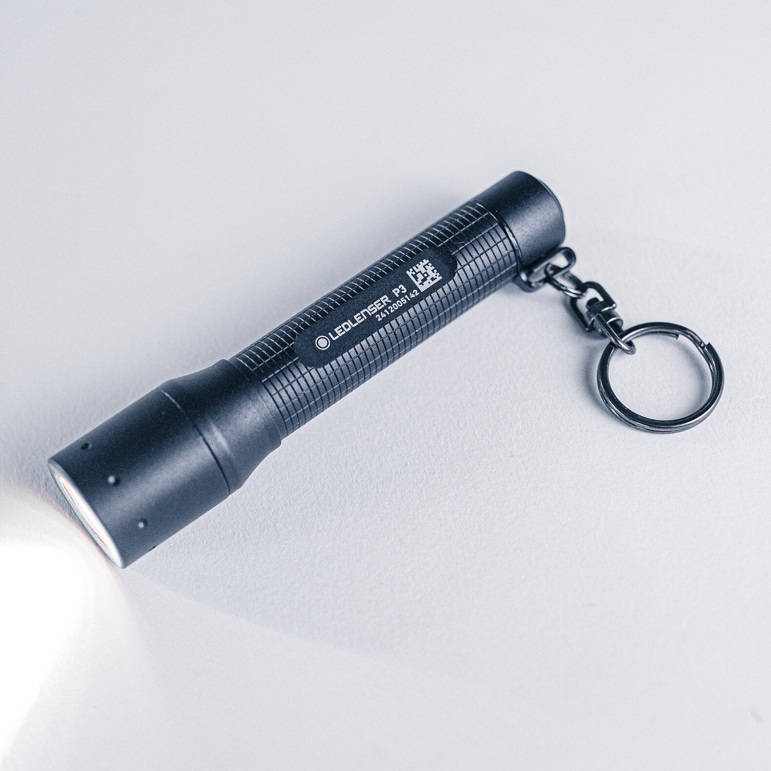 Eine schwarze LEDLENSER Ledlenser Taschenlampe P3 liegt auf einer hellen Oberfläche und strahlt einen weichen weißen Lichtstrahl aus. Die kompakte Taschenlampe hat einen strukturierten Griff und verfügt über einen Metall-Schlüsselring an einem Ende.