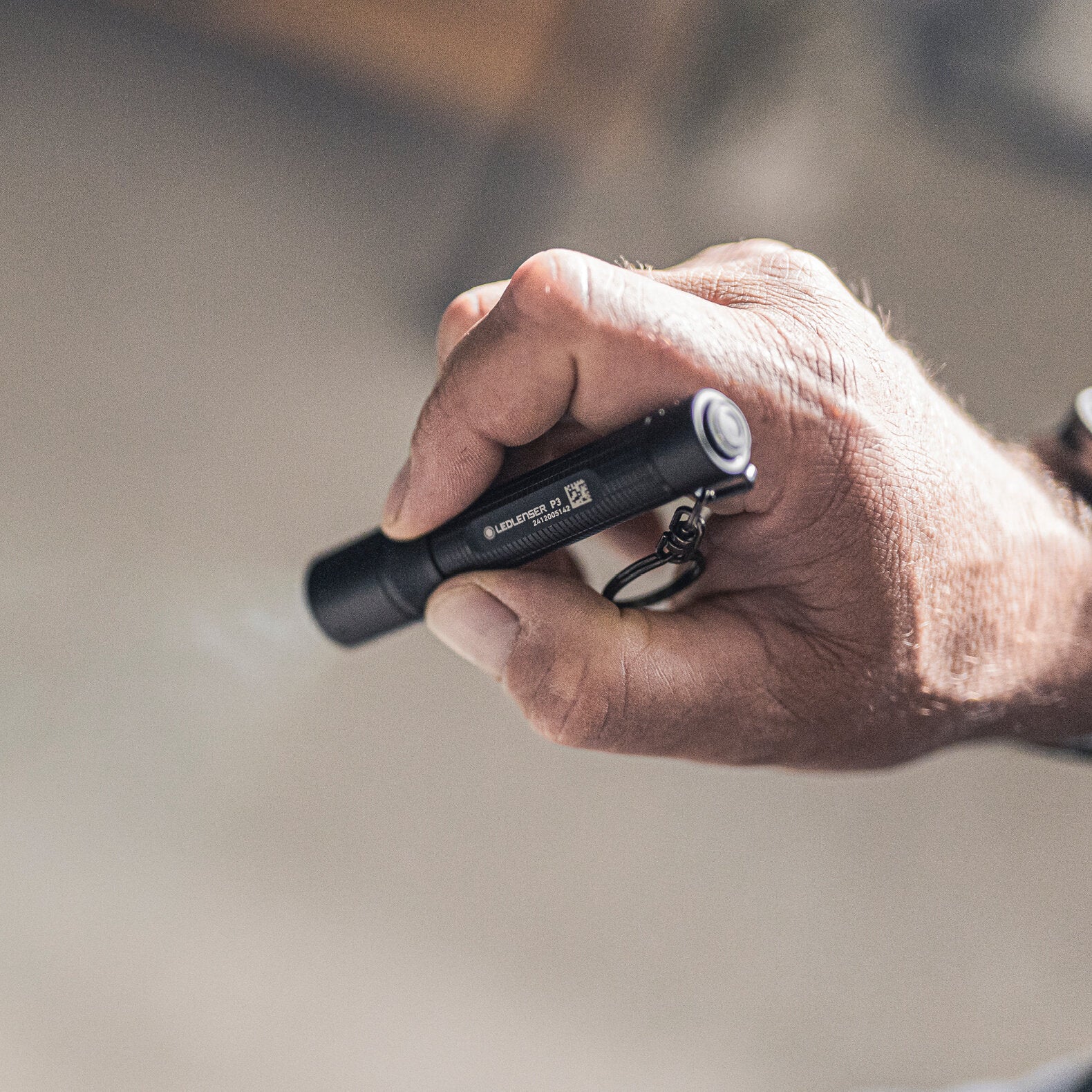 Eine Nahaufnahme zeigt eine Hand, die die LEDLENSER Ledlenser Taschenlampe P3 mit einem Schlüsselanhänger hält. Die kompakte Taschenlampe ist ausgeschaltet, der Hintergrund ist sanft verschwommen.