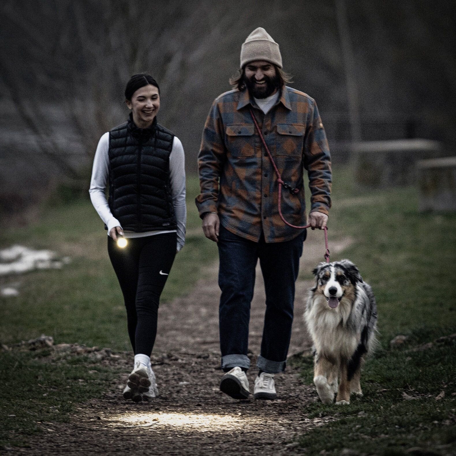 Eine Frau geht mit einem Mann auf einem Feldweg in der Dämmerung mit ihrem Hund spazieren. Sie trägt die LEDLENSER Ledlenser Taschenlampe P7R, während er, mit einer Mütze, die Leine hält. Beide lächeln und sind leger gekleidet für die freie Natur.