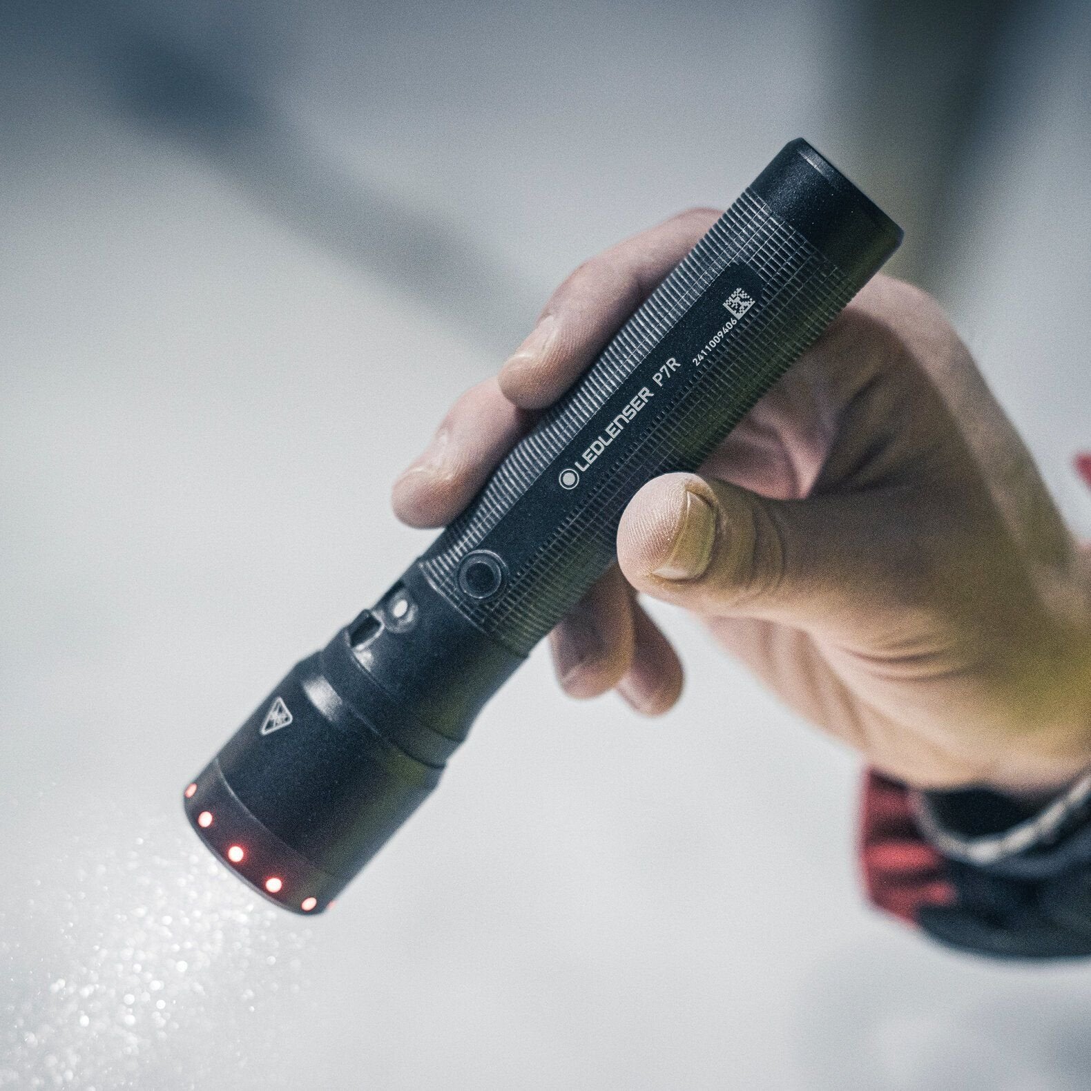 Eine Person hält eine LEDLENSER Ledlenser Taschenlampe P7R in der Hand, die eingeschaltet ist und einen Lichtstrahl von 2000 Lumen auf eine weiße Fläche projiziert. Die schwarze Taschenlampe hat einen strukturierten Griff und kleine rote Kontrollleuchten neben der Linse.