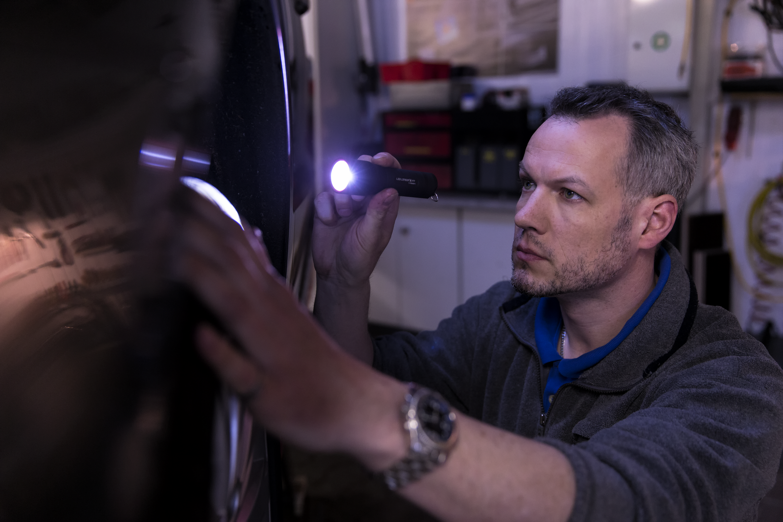 Ein Mann mit grauer Jacke und Armbanduhr inspiziert mit der LEDLENSER Taschenlampe P7 ein Fahrzeug in einer Werkstatt, umgeben von Werkzeugen. Das Advanced Focus System sorgt für präzise Beleuchtung bei der Detailarbeit.