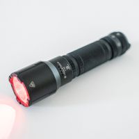 Eine schwarze LEDLENSER Ledlenser Taschenlampe TT3R ruht auf einer weißen Fläche, deren Linse rot leuchtet, um anzuzeigen, dass sie eingeschaltet ist und helles rotes Licht ausstrahlt.