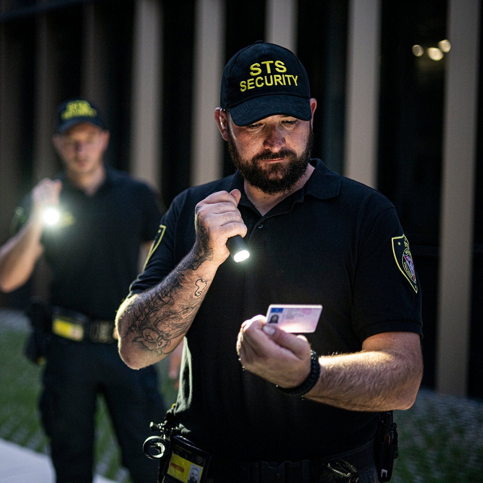 Zwei Sicherheitsbeamte in schwarzen Uniformen und Mützen mit STS Security-Logos überprüfen nachts im Freien einen Ausweis mit LEDLENSER Ledlenser Taschenlampe TT3R. Einer prüft den Ausweis genau, während der andere hinter ihm steht und ebenfalls eine Taschenlampe hält.