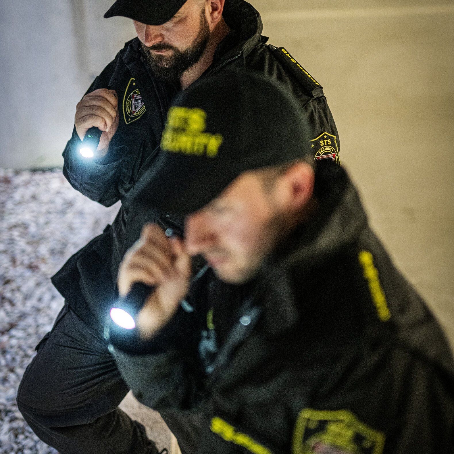 Nachts patrouillieren zwei Sicherheitskräfte mit SECURITY-Mützen in schwarzen Uniformen, die jeweils eine LEDLENSER TT3R Taschenlampe (1900 Lumen) tragen. Das gedämpfte Licht hebt die Sicherheitsaufnäher auf ihren Jacken hervor.