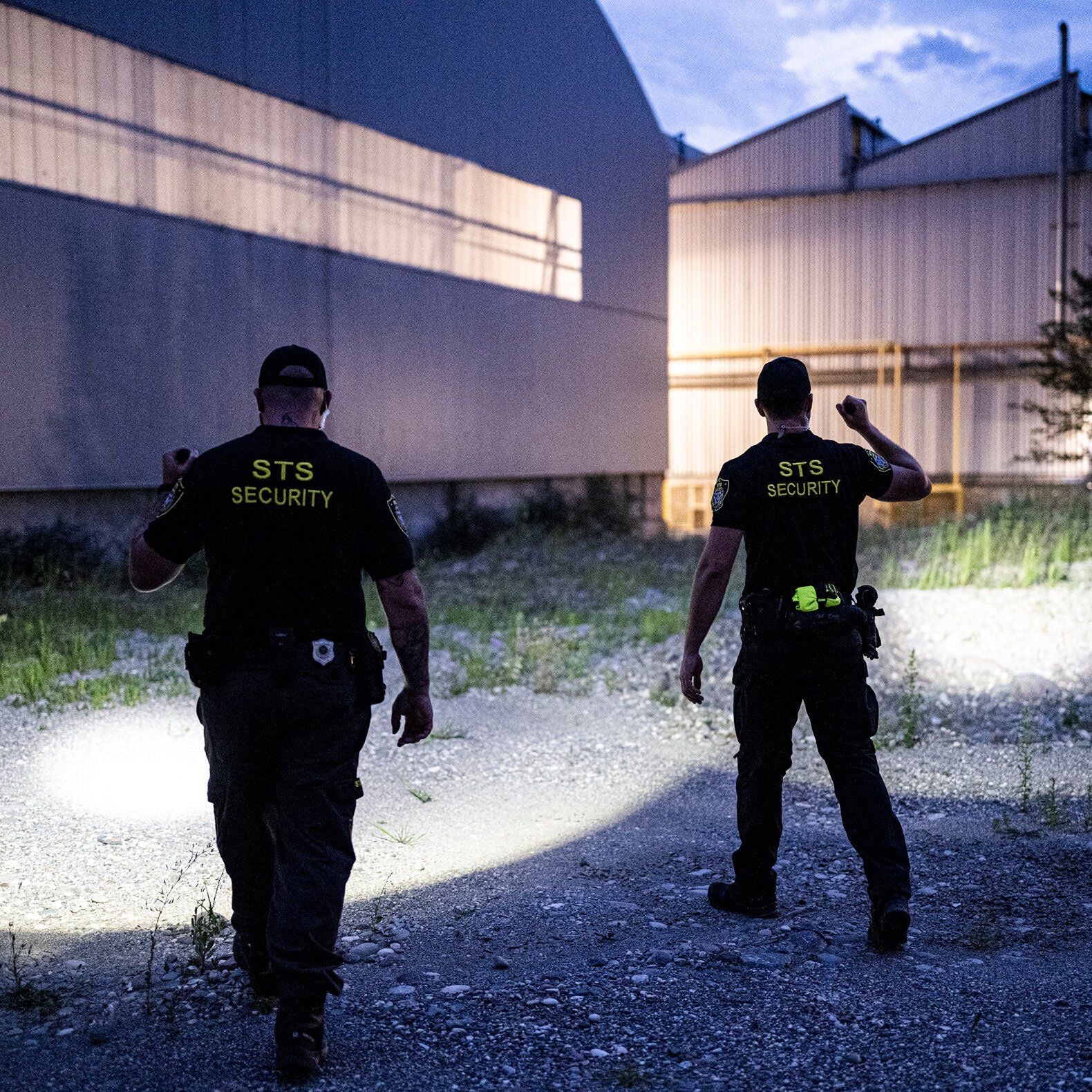 Zwei Sicherheitsleute in "STS Security"-Hemden patrouillieren in der Dämmerung durch ein Industriegebiet und beleuchten den Schotter in der Nähe großer Metallgebäude mit einer LEDLENSER Ledlenser Taschenlampe TT3R, die eine beeindruckende Lichtleistung von 1900 Lumen aufweist.