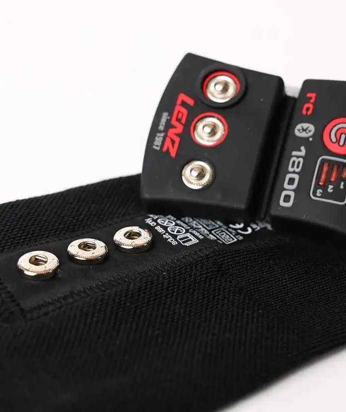 Nahaufnahme einer schwarzen LENZ Heizsocken 6.1 Toe Cap Merino Compression mit rotem und weißem Text, Metallschnappverschlüssen an einem Stoffband mit Pflegesymbolen - konzipiert für die Verwendung mit der Heat Socks Technologie.
