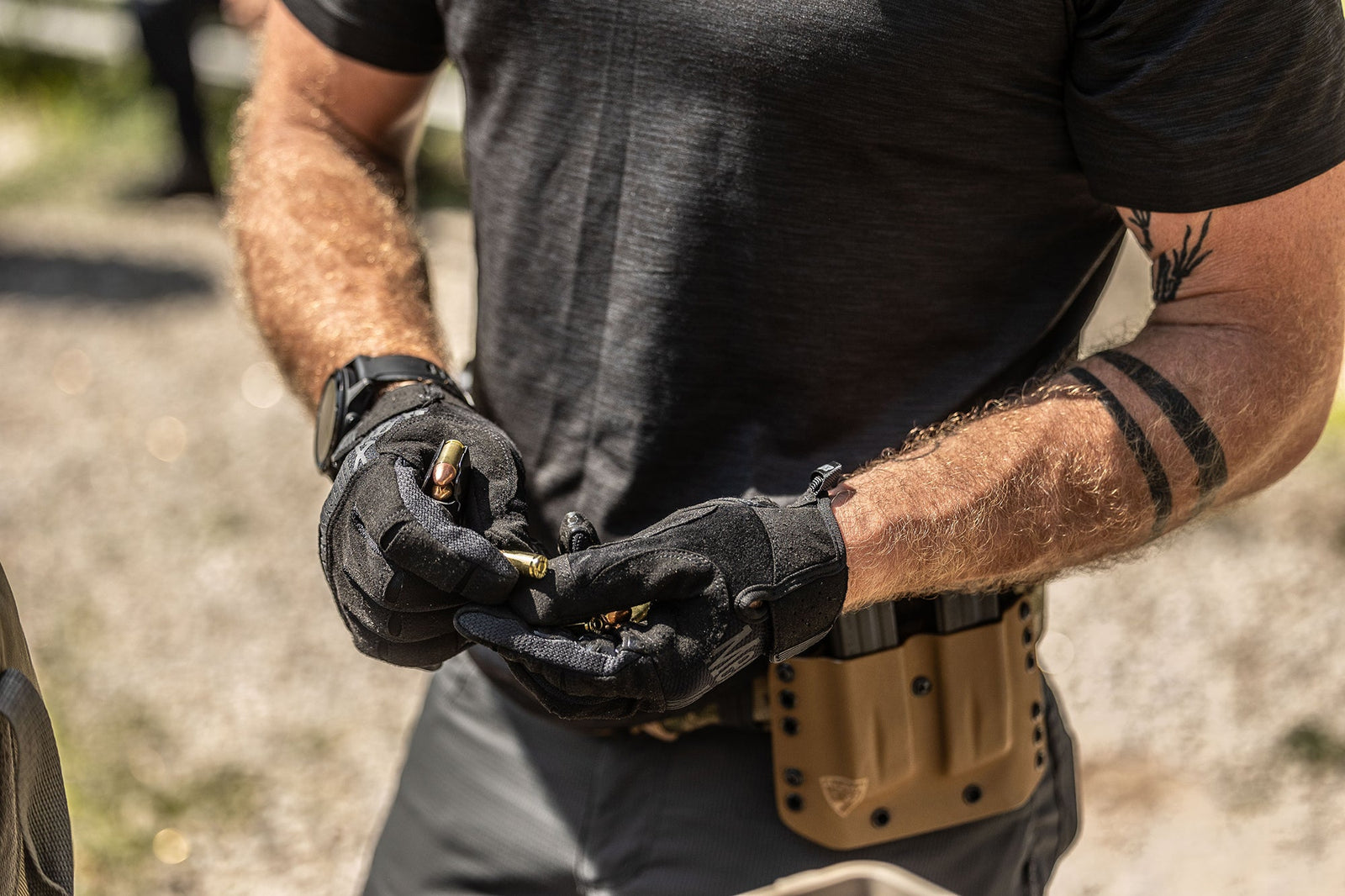 Eine Person im Freien mit taktischer Ausrüstung und einem Hüftholster trägt MECHANIX Mechanix Handschuhe Precision Pro - Covert Handschuhe und zeigt ein geometrisches Armbandtattoo auf dem Unterarm, während sie Kugeln hält.