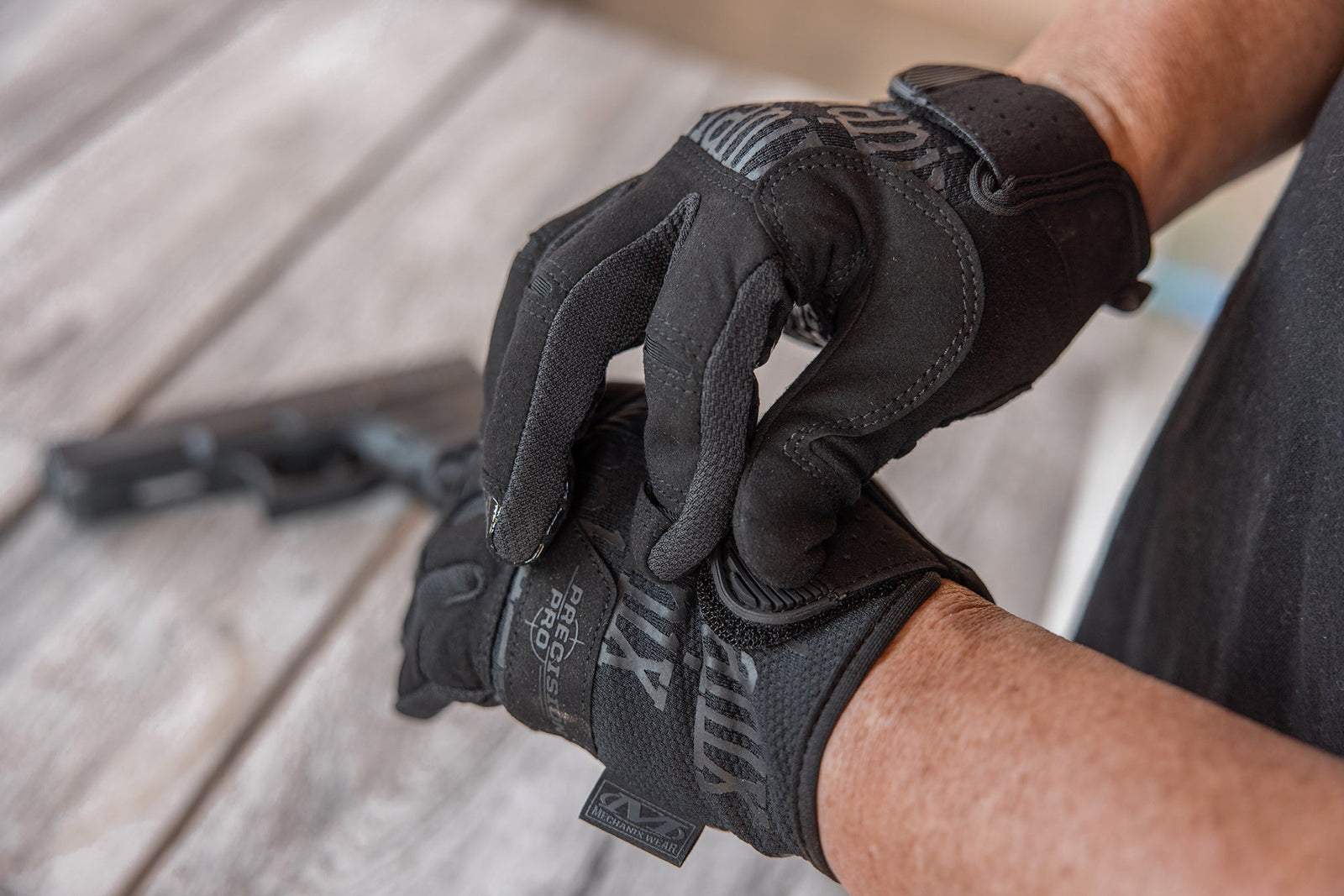 Eine Person, die MECHANIX Handschuhe Precision Pro - Covert trägt, stellt einen Handschuhriemen ein. Im unscharfen Hintergrund ruht eine Handfeuerwaffe auf verwittertem Holz, was die hohe Griffigkeit und Kontrolle der Handschuhe unterstreicht.