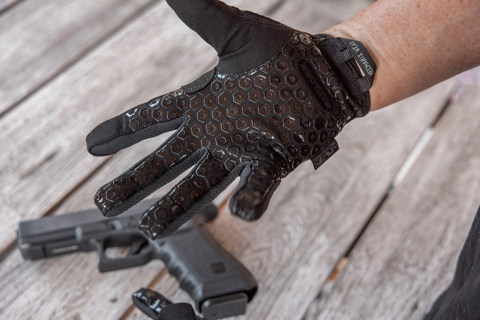 Eine behandschuhte Hand mit den MECHANIX Handschuhen Precision Pro - Covert mit schwarzem Sechseckmuster und hoher Fingerfertigkeit ist offen über einer hölzernen Oberfläche mit einer schwarzen Handfeuerwaffe auf dem Tisch im Hintergrund zu sehen.