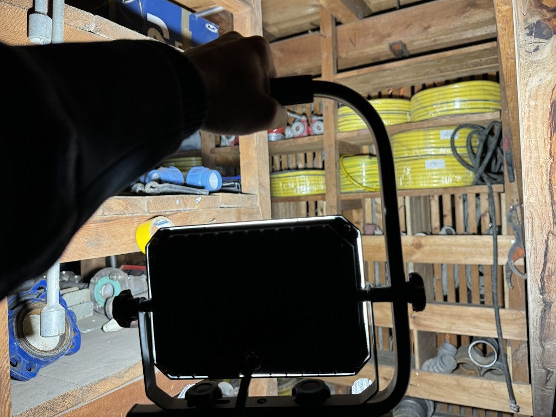 Eine Person benutzt einen NORDRIDE Nordride Baustrahler Sector 30W IP65 LED-Arbeitsscheinwerfer in einem Holzlager und richtet ihn auf Regale mit gelben Schläuchen, Rohren und Werkzeugen, um die Gegenstände deutlich auszuleuchten.