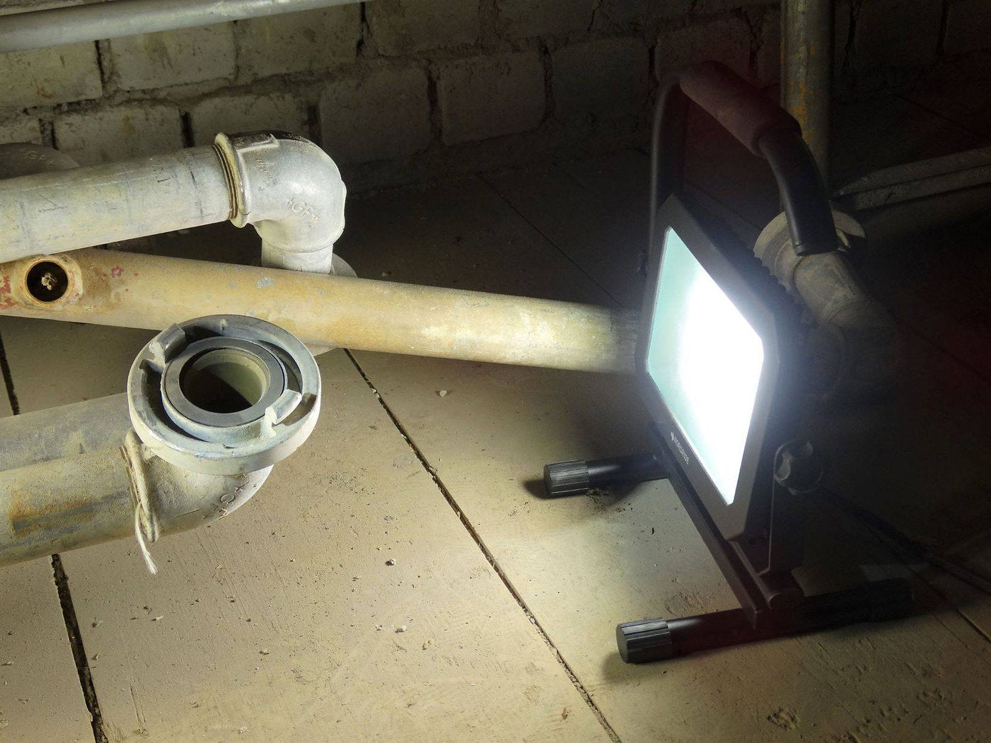 Ein Nordride Baustrahler Sector 50W IP65 von NORDRIDE leuchtet alte Metallrohre und einen Sperrholzboden in der Nähe einer Backsteinmauer in einem Keller oder Hauswirtschaftsraum hell aus.
