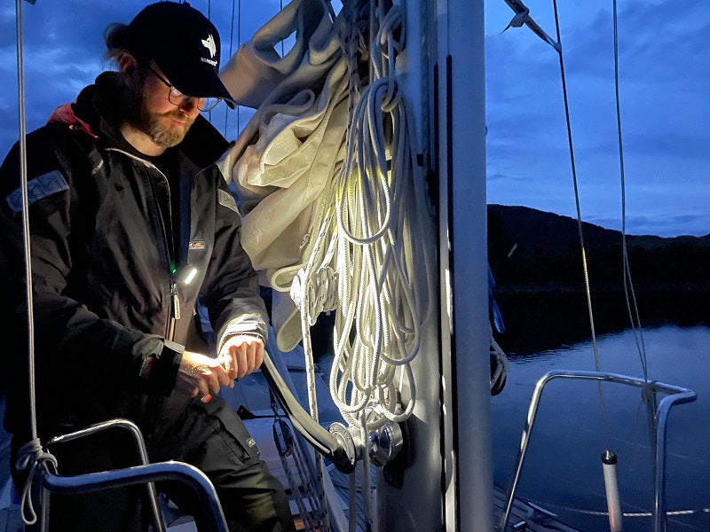 Eine Person in dunkler Jacke und Mütze arbeitet in der Dämmerung an einem Segelboot, das von einer NORDRIDE Nordride Clipleuchte Scout Red beleuchtet wird, wobei Taue und Segel sichtbar sind und das Wasser im Hintergrund ruhig ist.