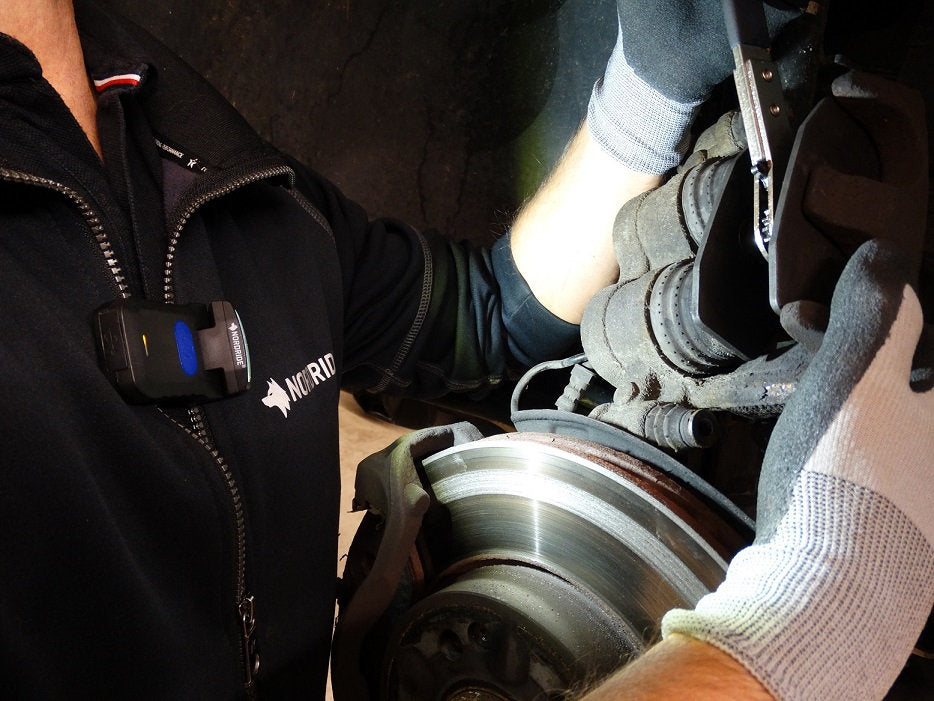 Ein Mechaniker in Handschuhen und dunkler Jacke stellt den Bremssattel und die Bremsscheibe eines Autos ein. Er arbeitet unter der hellen Beleuchtung der NORDRIDE Nordride Clipleuchte Scout Red.