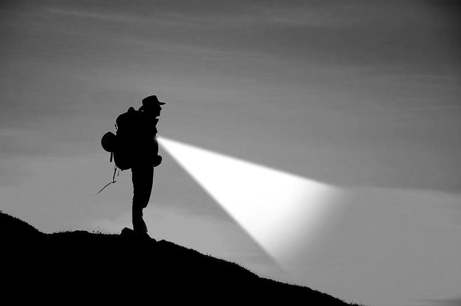 Eine Silhouette mit Hut und Rucksack steht nachts auf einem Hügel und leuchtet mit einer Nordride Clipleuchte Scout Red von NORDRIDE nach vorne, um den Boden vor sich zu beleuchten. Die schwarz-weiße Szene hebt den Strahl der Lampe hervor.