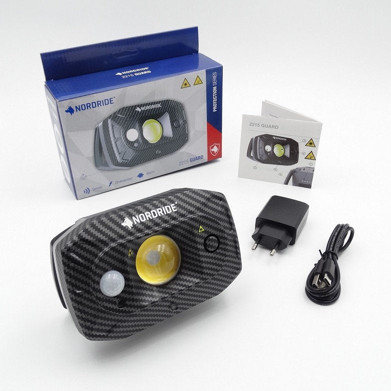Der NORDRIDE Flutlichtstrahler Guard überzeugt mit Carbonfaser-Optik, IP67-Zertifizierung und umfangreichen Sicherheitsfunktionen. Im Lieferumfang enthalten: Box, Ladeadapter, USB-Kabel und Anleitung auf weißem Hintergrund.