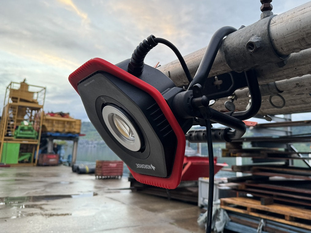 Der NORDRIDE Hochleistungsstrahler Lumos 100 W, eine tragbare LED-Leuchte mit 10.000 Lumen und einem rot-schwarzen Gehäuse, ist im Freien an ein Metallrohr geklemmt. Im Hintergrund sind Baugeräte und gestapeltes Material auf nassem Beton zu sehen.