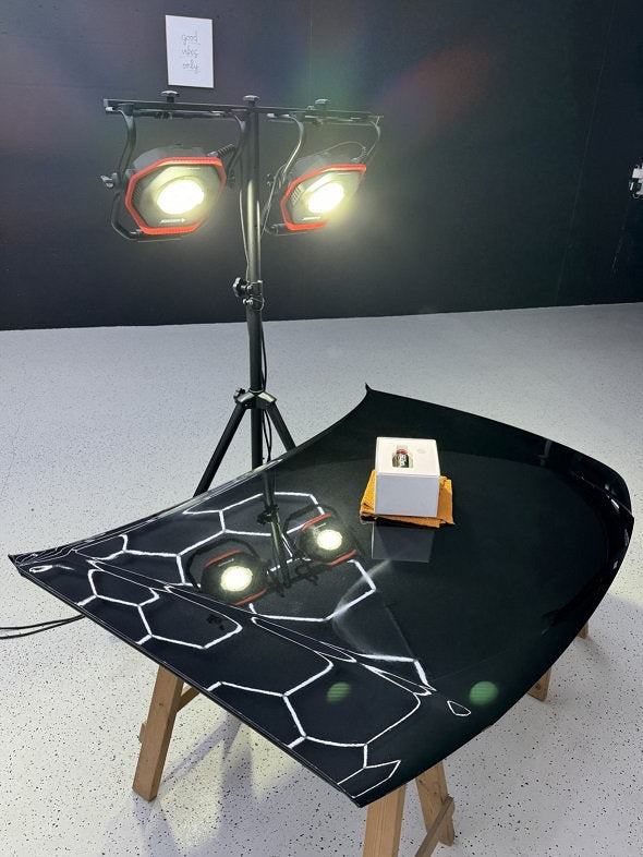 Zwei NORDRIDE Nordride Hochleistungsstrahler Lumos 100 W mit je 10.000 Lumen beleuchten hell eine schwarz glänzende Autohaube auf Holzständern. Die Motorhaube reflektiert die Lichter und Muster. Darauf steht eine kleine weiße Box mit einer Flasche.