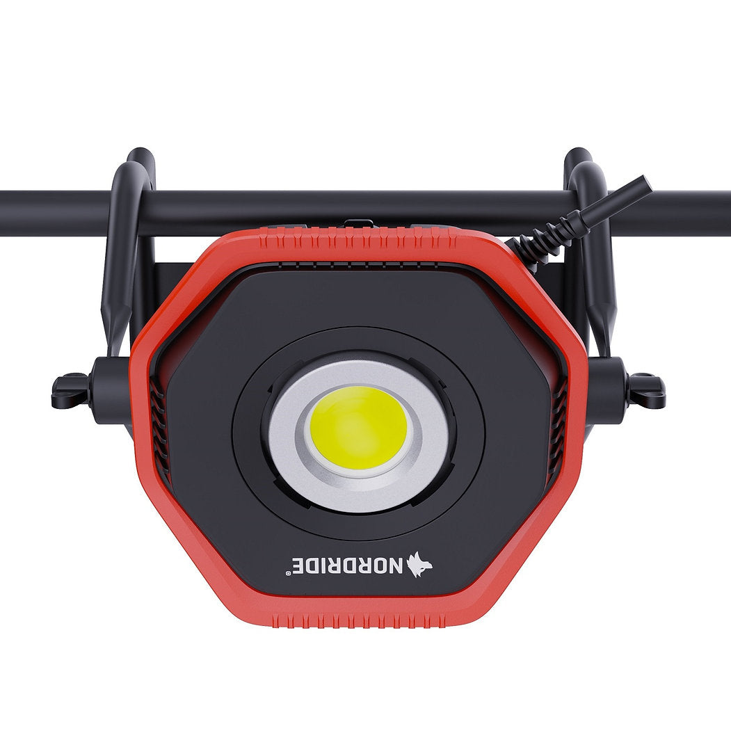Der NORDRIDE Hochleistungsstrahler Lumos 100 W ist ein rot-schwarzer, sechseckiger LED-Arbeitsscheinwerfer mit Wolfslogo, der an einer horizontalen Stange montiert ist, über ein Stromkabel rechts oben verfügt und beeindruckende 10.000 Lumen für jede Baustelle liefert.