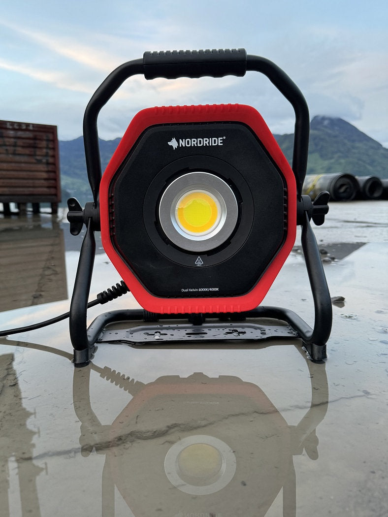 Ein tragbarer NORDRIDE Nordride Hochleistungsstrahler Lumos 100 W mit einem rot-schwarzen Rahmen steht auf nassem Grund, seine Reflexion ist im Wasser sichtbar, während Berge und industrielle Strukturen im Hintergrund verschwimmen.