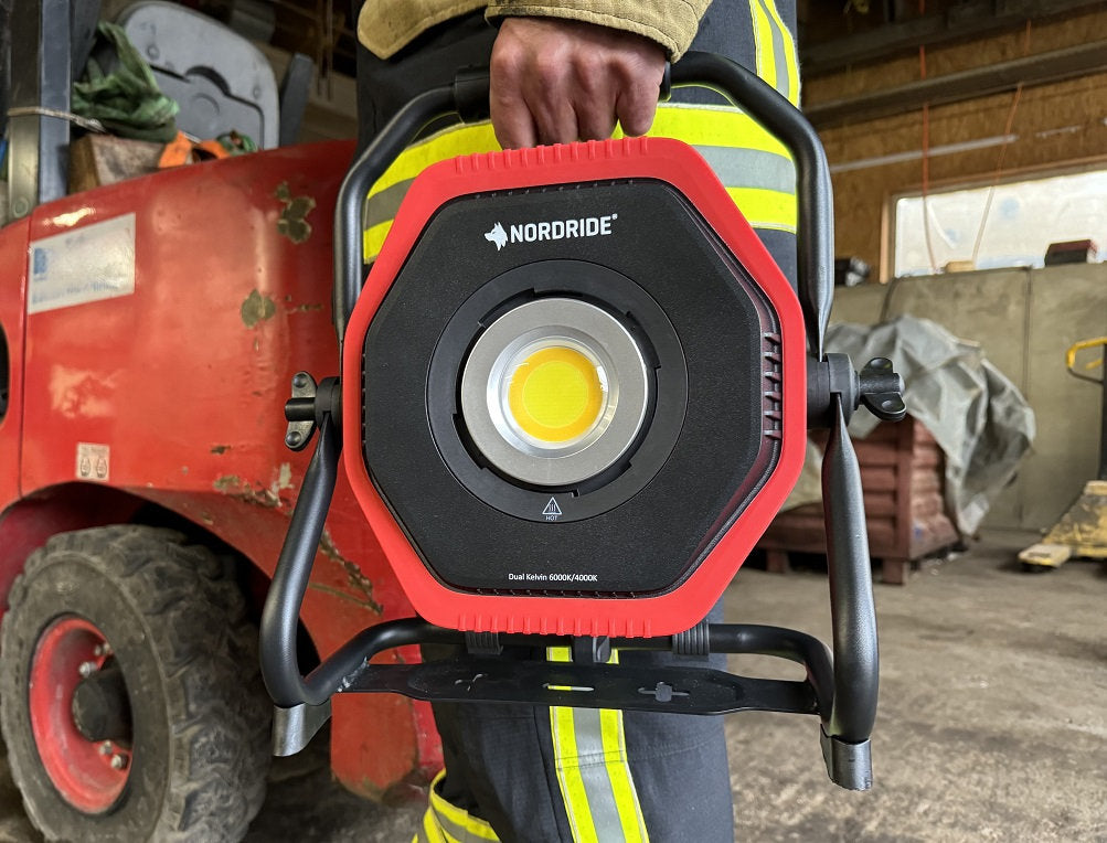 Eine Person in einer gelb gestreiften Arbeitshose hält einen rot-schwarzen NORDRIDE Nordride Hochleistungsstrahler Lumos 80W R Dual Kelvin in einer industriellen Umgebung, in der Maschinen und Materialien im Hintergrund zu sehen sind.
