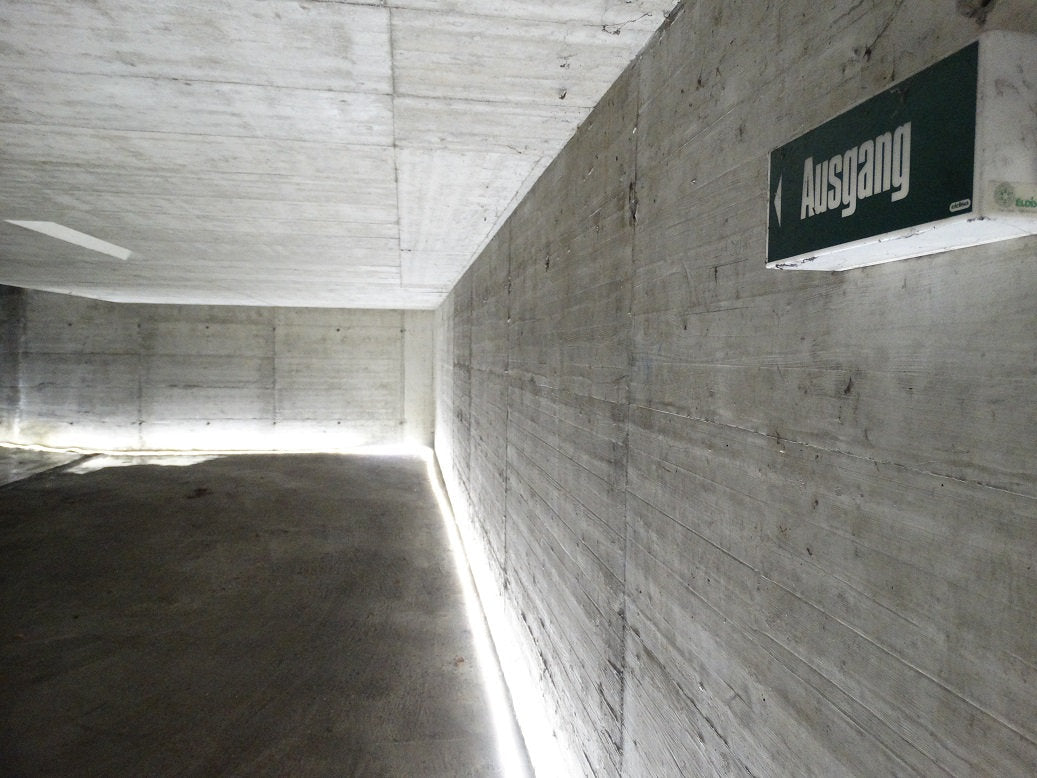 In einem Betonflur mit minimaler Beleuchtung befindet sich an der rechten Wand ein grünes "Ausgang"-Schild, das nach links zeigt. Für eine dezente Beleuchtung sorgt das NORDRIDE Nordride Lichtband Viper LED 25m, das die Sichtbarkeit des Flurs verbessert.