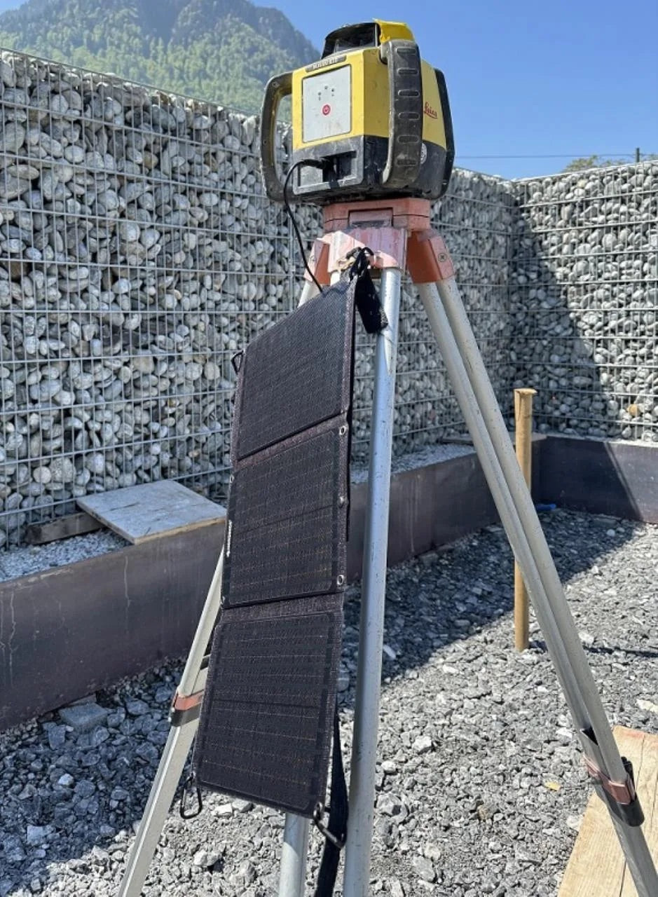 Ein Stativ hält eine gelb-schwarze Laserwasserwaage, an deren Bein ein NORDRIDE SOLAR PULSE 24W-Solarpanel befestigt ist. Die Anlage steht auf Kies, umgeben von Maschendraht-Steinmauern und Bergen im Hintergrund.