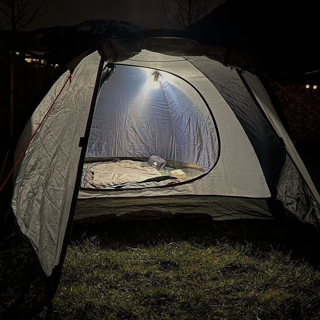 Ein Zelt leuchtet in der Nacht auf einem grasbewachsenen Boden, dessen geöffnete Klappe eine gemütliche Einrichtung mit Decken und Kissen um eine hängende NORDRIDE Signalkappe und einen Diffusor für Pfadfinder-, Patrouillen- und Sanitäterlicht offenbart. Im Hintergrund sind Bäume und Berge schwach zu erkennen.