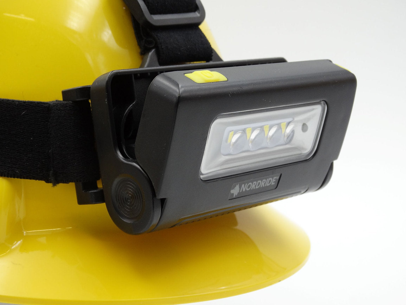 Nahaufnahme eines gelben Schutzhelms, an dem die NORDRIDE Nordride Strinlampe Duo befestigt ist, mit einer rechteckigen LED-Arbeitslampe und einem gelben Einschaltknopf auf der Oberseite.