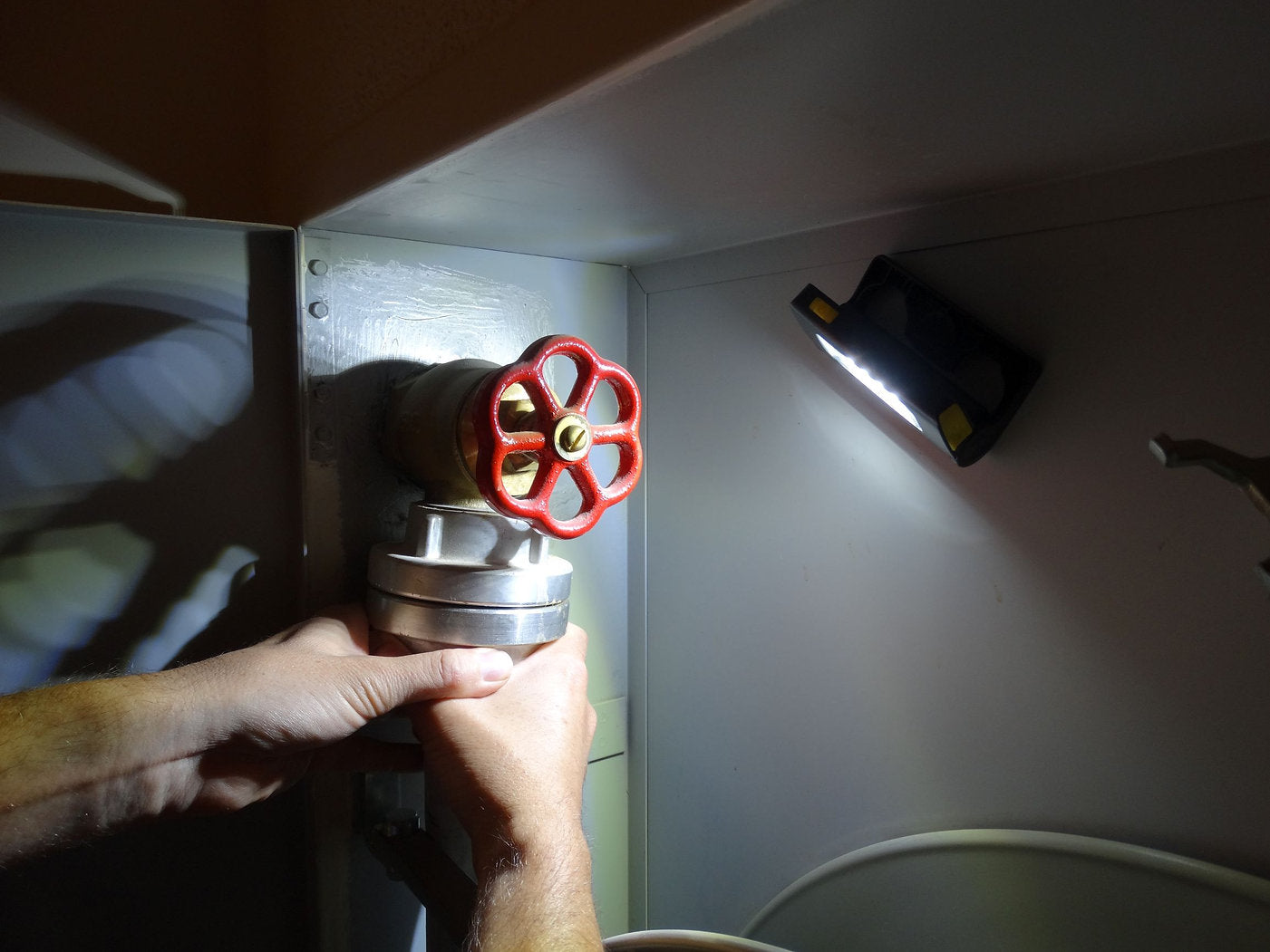 Die Hände einer Person drehen ein großes rotes Ventil in einem Metallschrank, das von einer an der Schrankwand montierten NORDRIDE Nordride Strinlampe Duo beleuchtet wird.