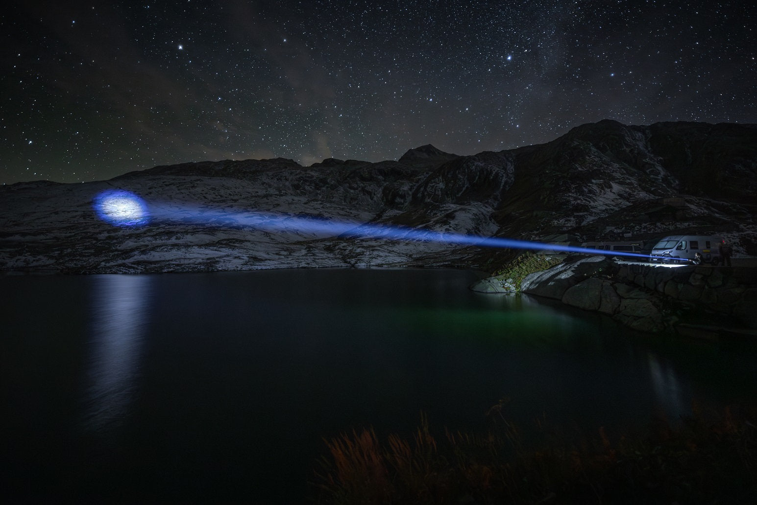 Eine Person steht neben einem Lieferwagen an einem nächtlichen See und leuchtet mit einer NORDRIDE Nordride Taschenlampe Spot Long Ranger R über das Wasser auf schneebedeckte Berge unter einem sternenklaren Himmel.