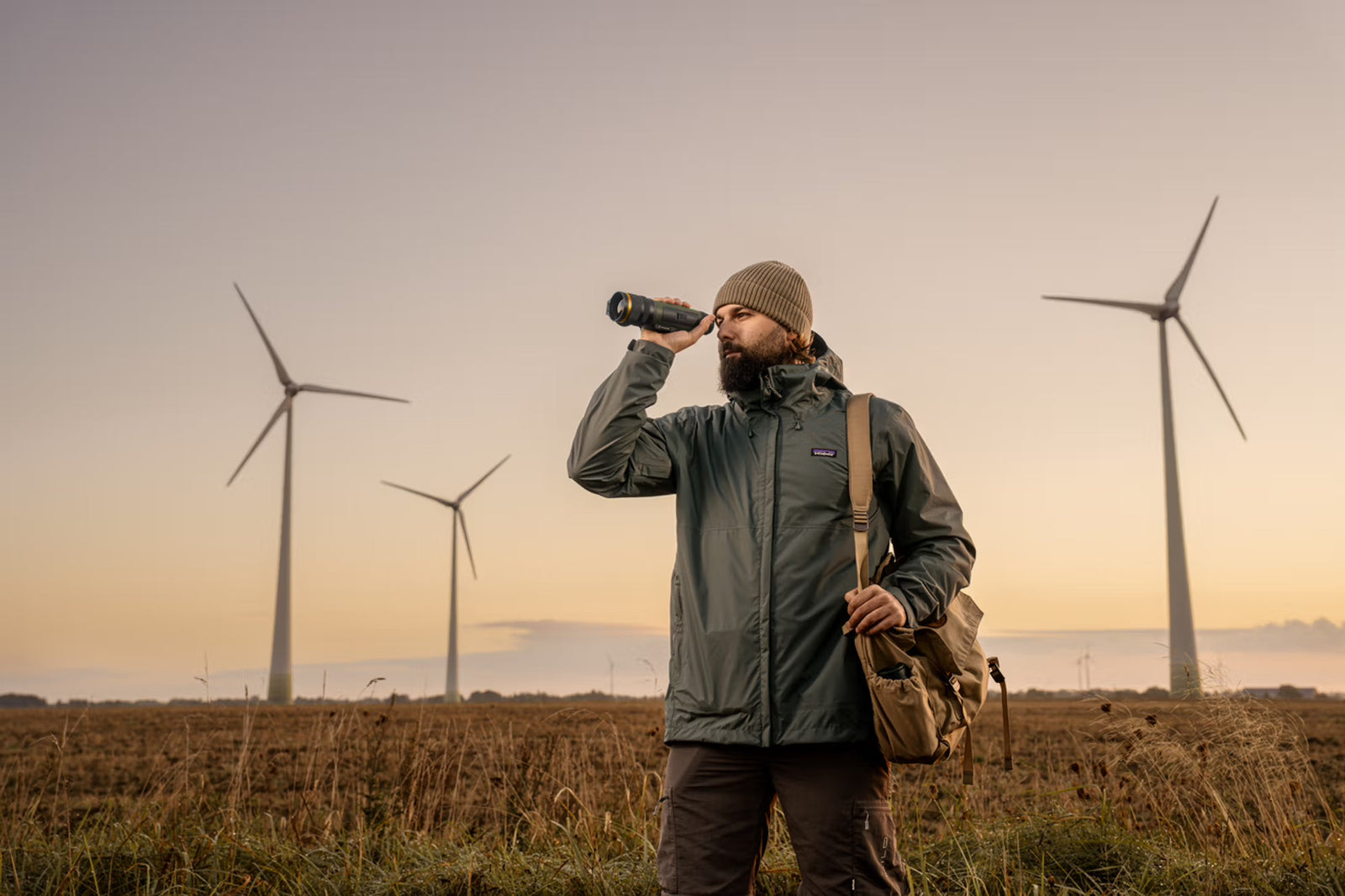Ein Mann in Outdoor-Kleidung mit Mütze und Umhängetasche steht bei Sonnenuntergang auf einem Feld und schaut durch sein PULSAR Pulsar WLO Wärmebildgerät Lumion XL50, während sich im Hintergrund mehrere Windräder erheben.