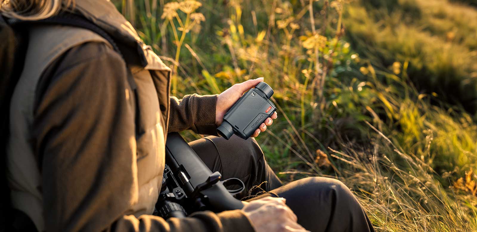 Eine Person sitzt im hohen Gras unter warmem Sonnenlicht, trägt Outdoor-Kleidung und hält ein PULSAR Pulsar Wärmebildgerät Axion XG35 Compact Fernglas in der einen und ein Gewehr in der anderen Hand.