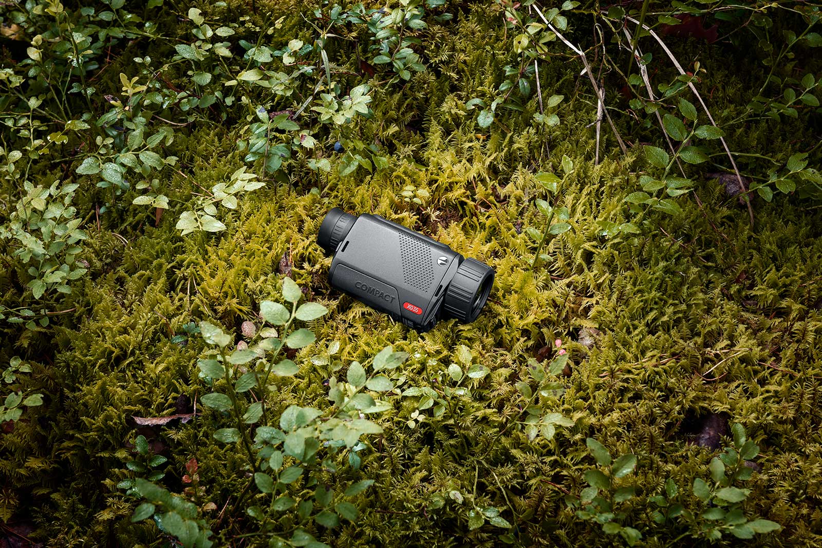 Ein PULSAR Pulsar Wärmebildgerät Axion XG35 Compact in schwarz mit rotem Knopf ruht auf grünem Moos zwischen kleinen Pflanzen und Blättern in einer Waldlandschaft.