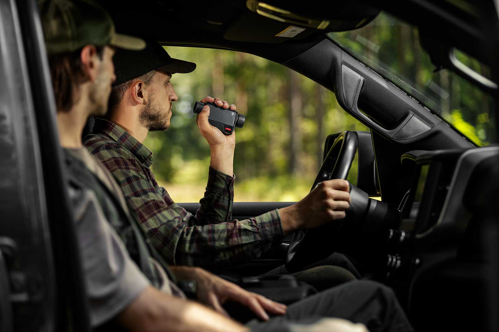 Zwei Männer sitzen in einem Fahrzeug; der Fahrer, der ein kariertes Hemd und eine Mütze trägt, benutzt ein PULSAR Pulsar Wärmebildgerät Axion XG35 Compact, um den Außenbereich zu scannen, während der Beifahrer inmitten von verschwommenem Grün nach vorne schaut.