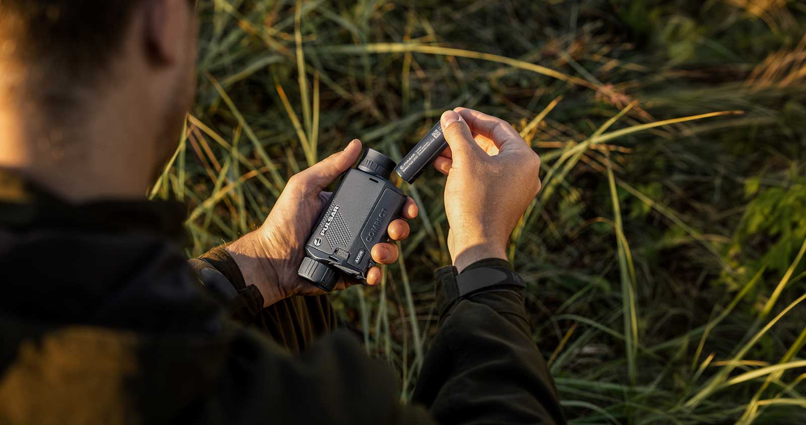 Im Freien setzt jemand eine Batterie in ein PULSAR Axion XG35 Compact Wärmebildgerät ein, während im Hintergrund grünes Gras zu sehen ist.